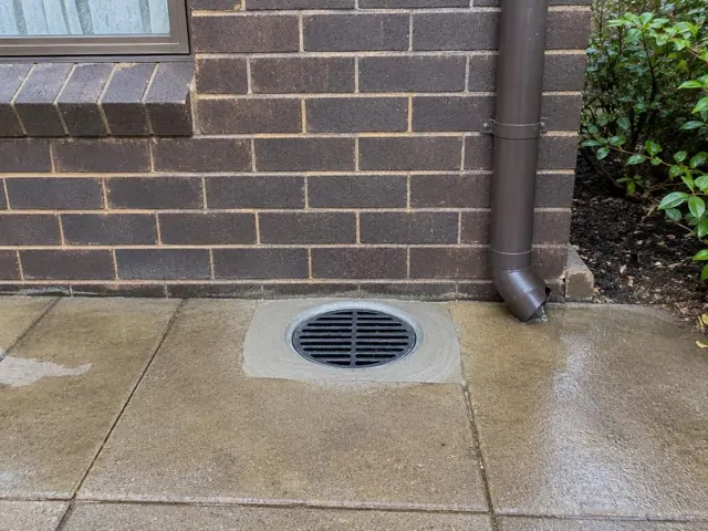 Clean round stormwater grate set in concrete paving beside a brick house wall in Oatlands