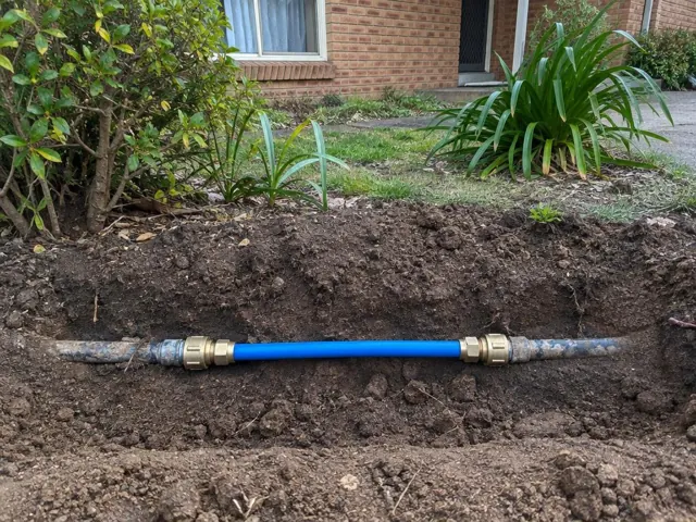 Repaired blue water pipe section in a dirt trench beside a brick house in Carlingford