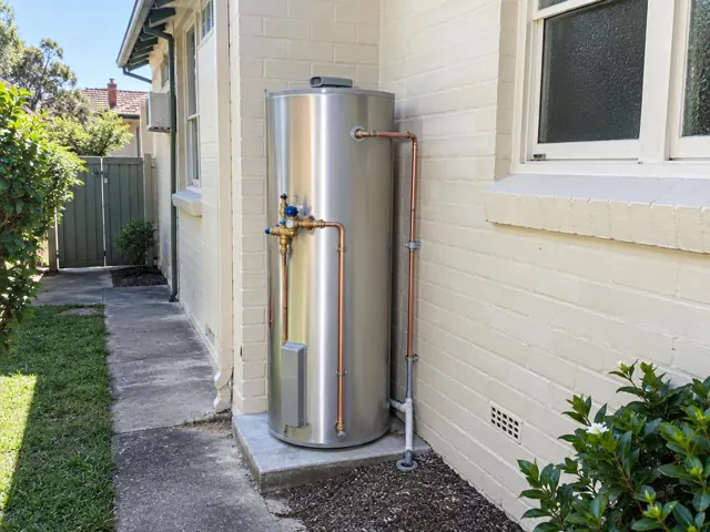 Outdoor stainless steel hot water cylinder beside a cream brick wall and window in Epping